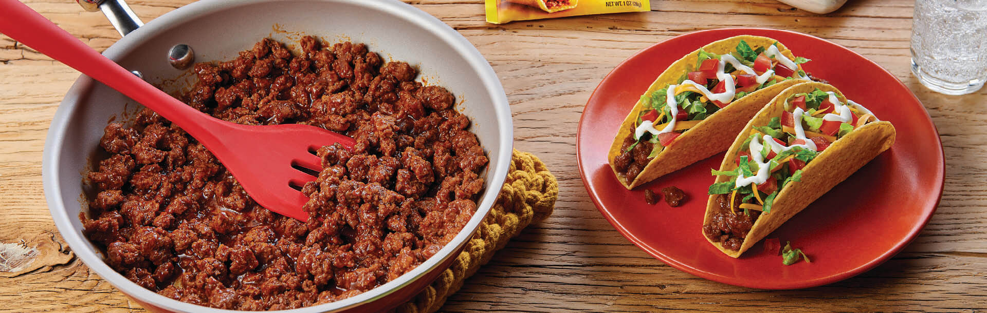 padella di carne macinata condita con una spatola rossa accanto a tacos duri e carichi su un piatto rosso